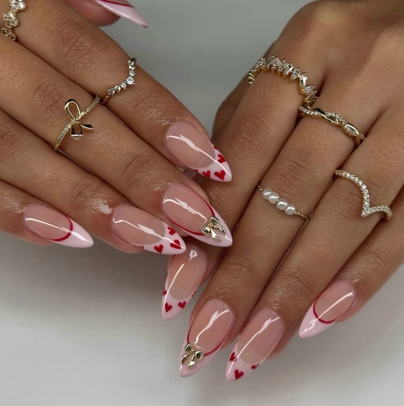 Close-up of hands with manicured nails featuring clear polish and red heart accents, adorned with multiple gold and silver rings.