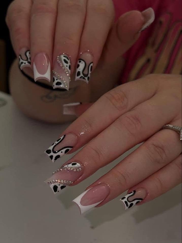 Close-up of manicured hands with long, coffin-shaped nails featuring cow print design and rhinestone accents.