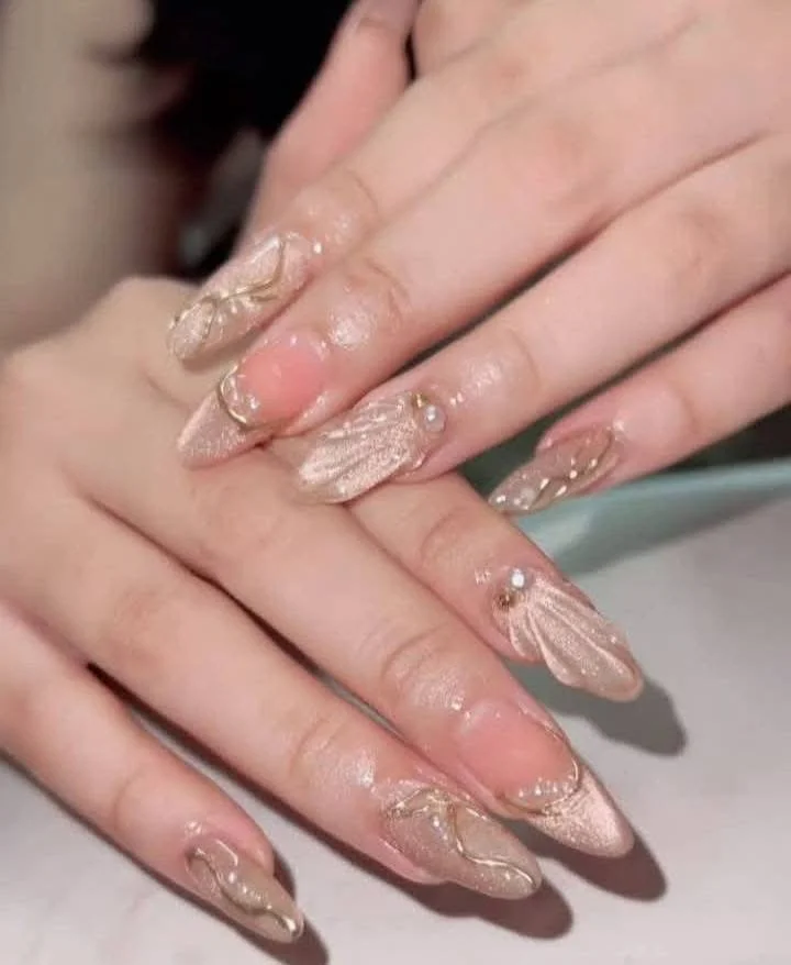 Close-up of hands with long, almond-shaped nails featuring a nude and gold marbled design with small pearl and gold embellishments.