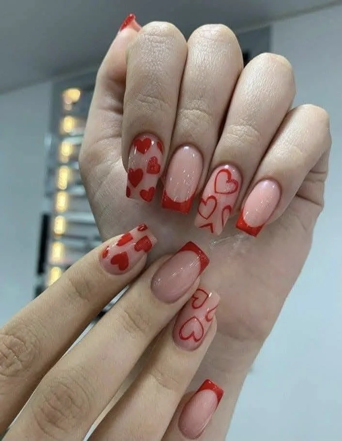 Close-up of hands with decorated nails featuring red and pink hearts, some nails with red French tips, and a soft pink base.