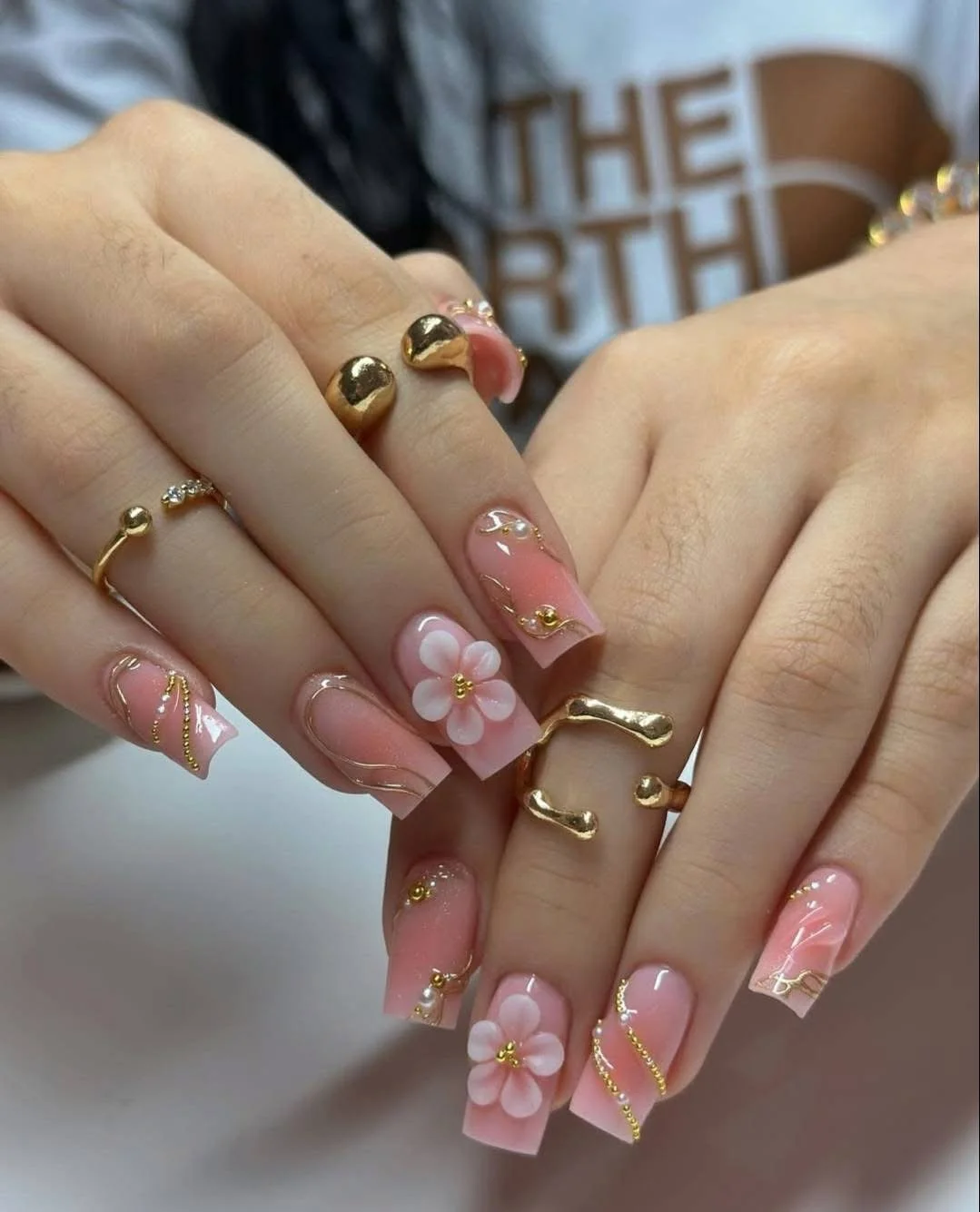 Close-up of hands with decorated nails and gold rings, featuring pink and white floral designs and gold accents.