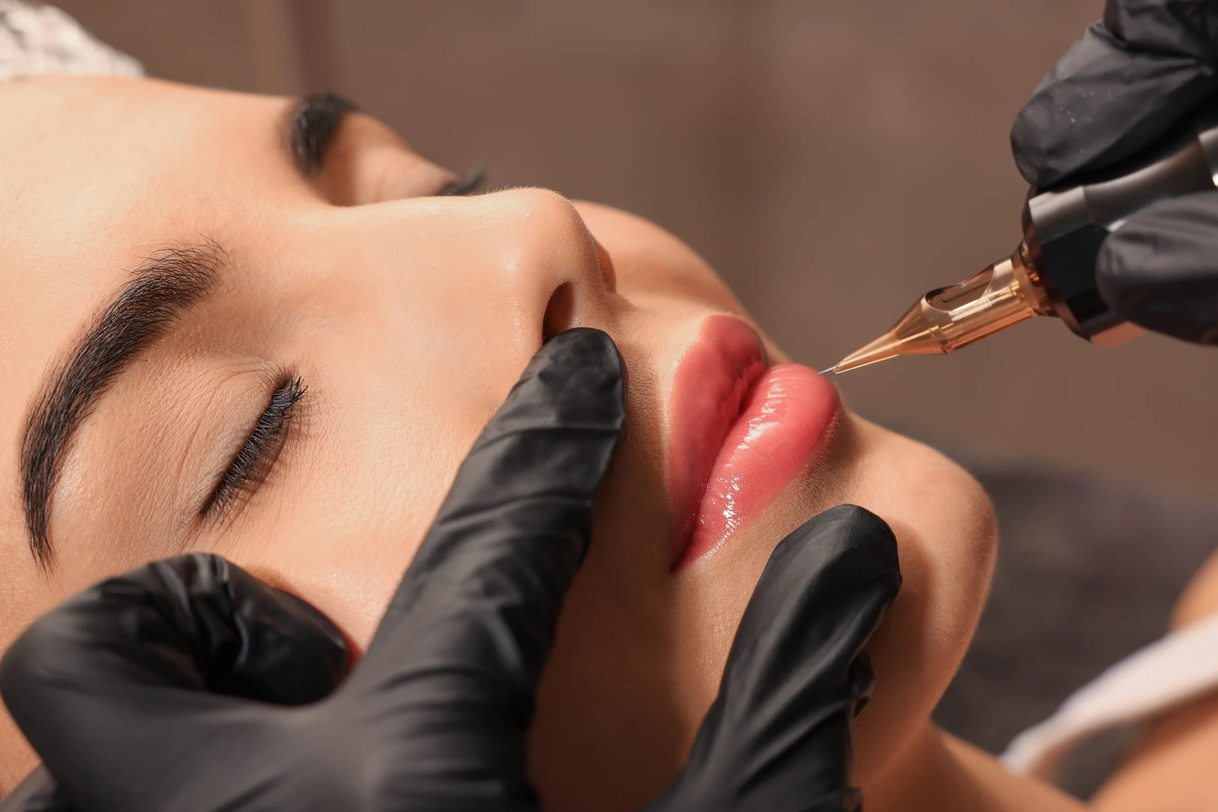 Close-up of a woman receiving a lip tattoo or permanent makeup procedure. The technician, wearing black gloves, is using a tattoo needle to apply pigment to the woman's lips, which are parted.