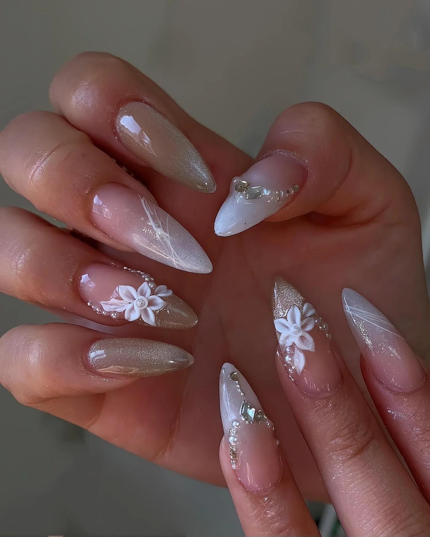 Close-up of a hand with long, almond-shaped nails decorated with white flowers, diamonds, and glitter, featuring a nude pink base color.