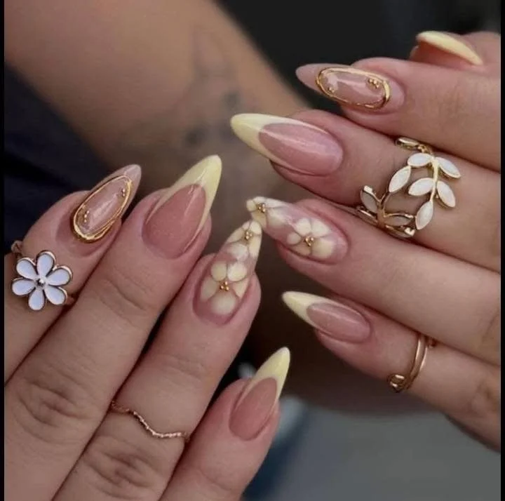 Close-up of hands with beautifully manicured nails in a pink and yellow gradient, adorned with decorative rings, including a white flower, gold leaves, and pearl accents.