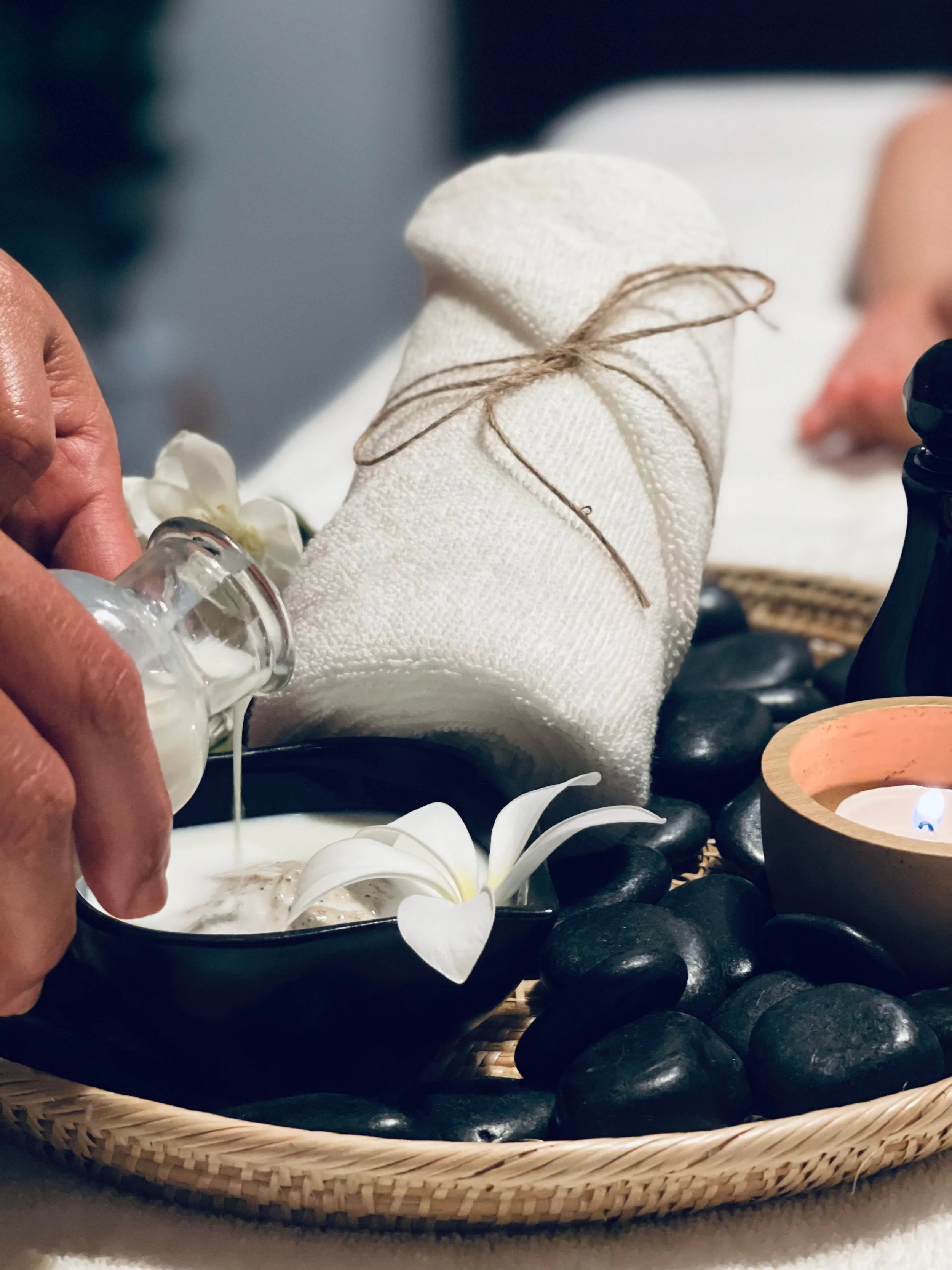 A person holds a small glass bottle of lotion or oil, near a black bowl with white flowers and candles, surrounded by black stones, on a woven tray.