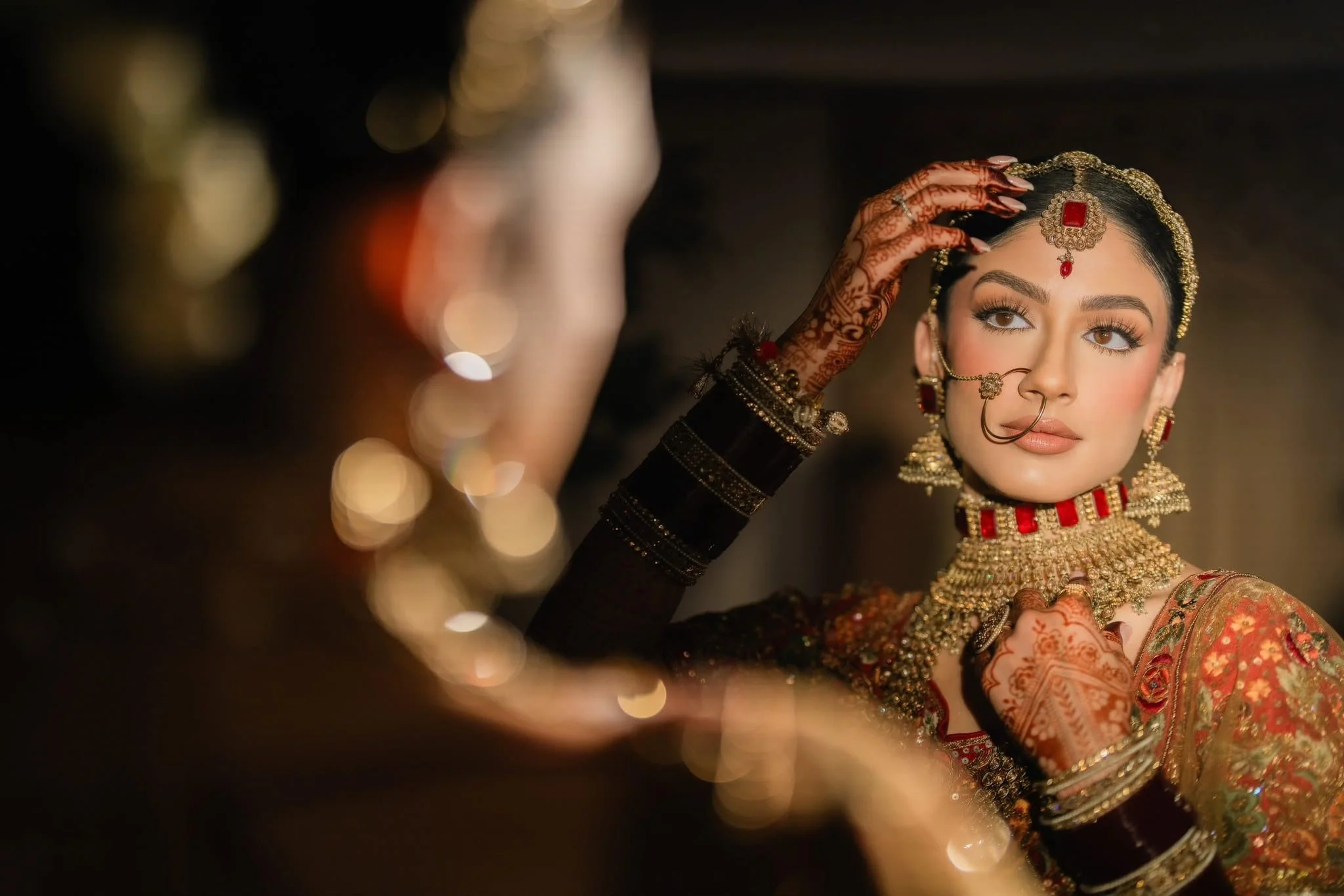 A woman dressed in traditional Indian bridal attire, wearing elaborate jewelry, including a maang tikka, nose ring, earrings, and necklace, adjusting her hair with Henna decorated hands.