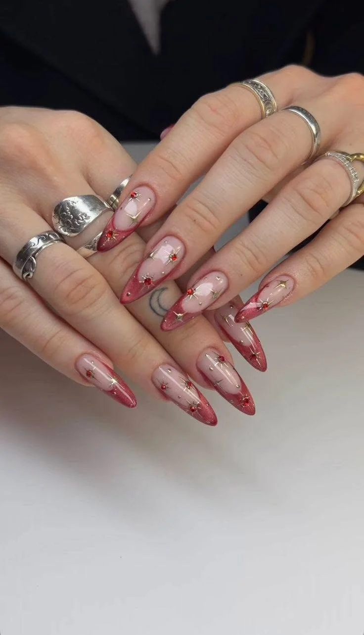 Close-up of a person's hands with long almond-shaped nails decorated with red, pink, and gold nail art, and wearing multiple silver rings.