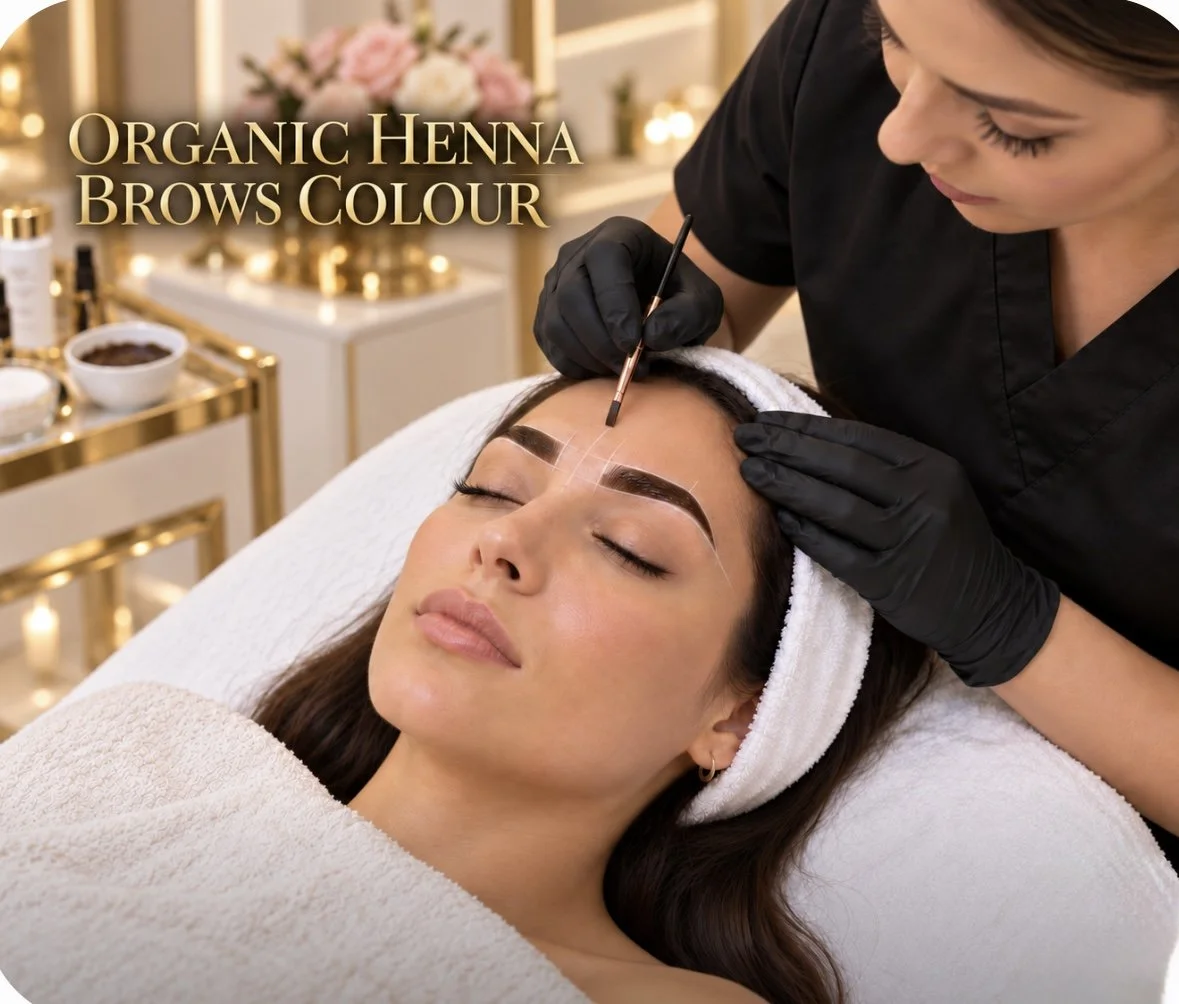 A woman receiving a henna eyebrow tinting treatment in a beauty salon, lying down with her eyes closed, a beauty professional applying dye to her eyebrows with a small brush, with a sign reading 'Organic Henna Brows Colour' in the background.