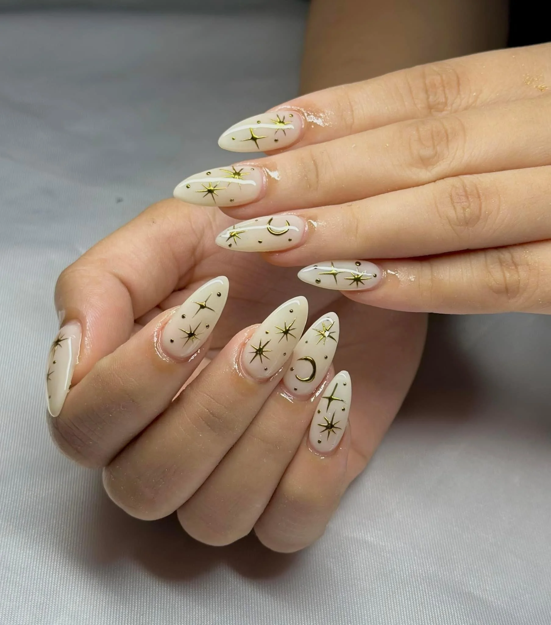 Close-up of manicured nails painted in a light beige color with gold star, moon, and dot designs.