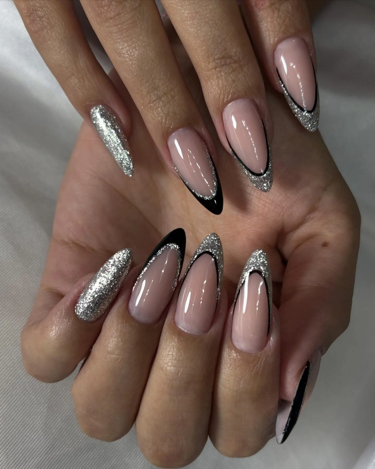 Close-up of manicured nails with a nude base, black tips, silver glitter accents, and a glittery silver thumb. The nails are almond-shaped.