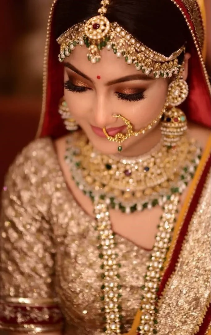 A bride dressed in traditional Indian bridal attire, wearing an ornate gold and pearl jewelry set including a headpiece, earrings, nose ring, and layered necklaces, with her eyes closed and smiling gently.