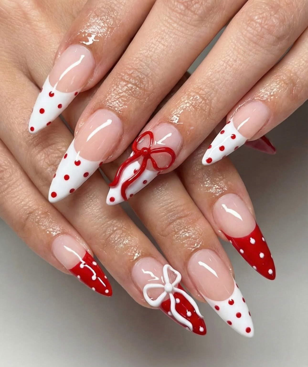 Close-up of hands with long, almond-shaped nails painted in a red and white polka dot pattern. The nails also feature three-dimensional white bows and red ribbon designs.
