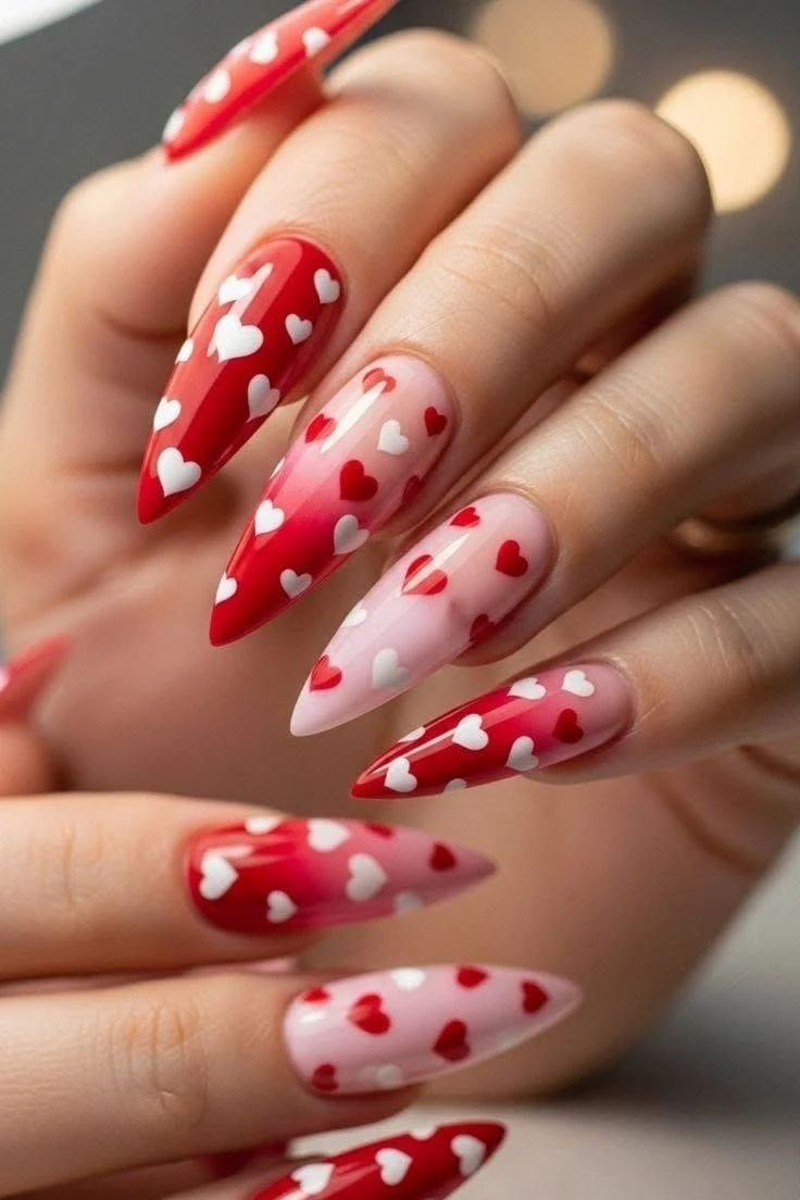 Close-up of a hand with long, pointed, pink and red acrylic nails decorated with small white and red hearts.