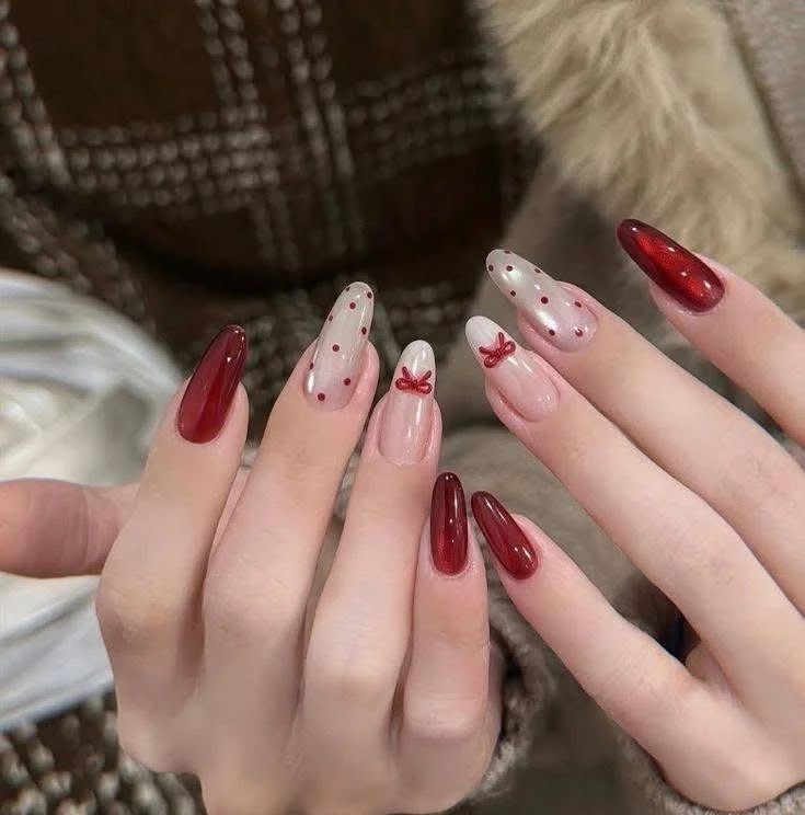 Close-up of a person's hands with red and clear nail polish, decorated with polka dots, bows, and a shiny finish.