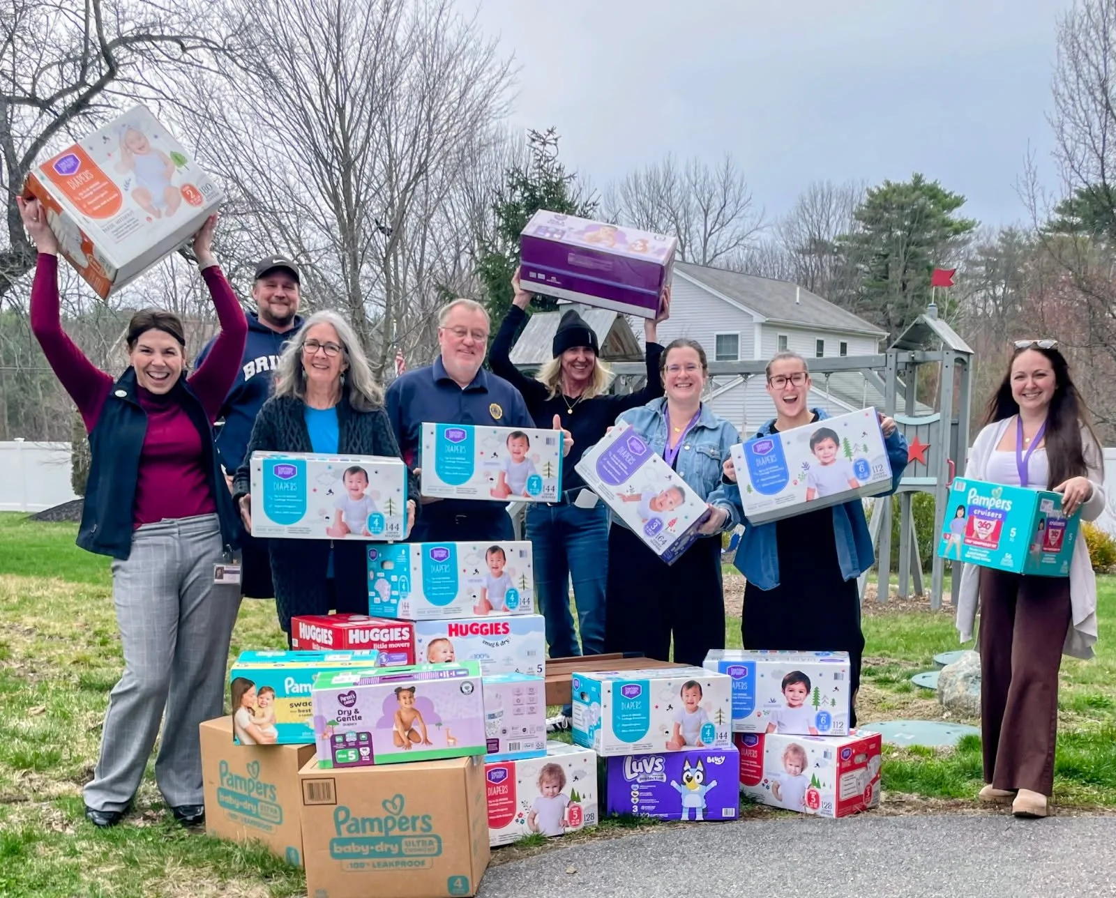 A group of people smiling and holding boxes of diapers and baby supplies outdoors. There are several large boxes of Pampers diapers and Huggies wipes in the foreground.