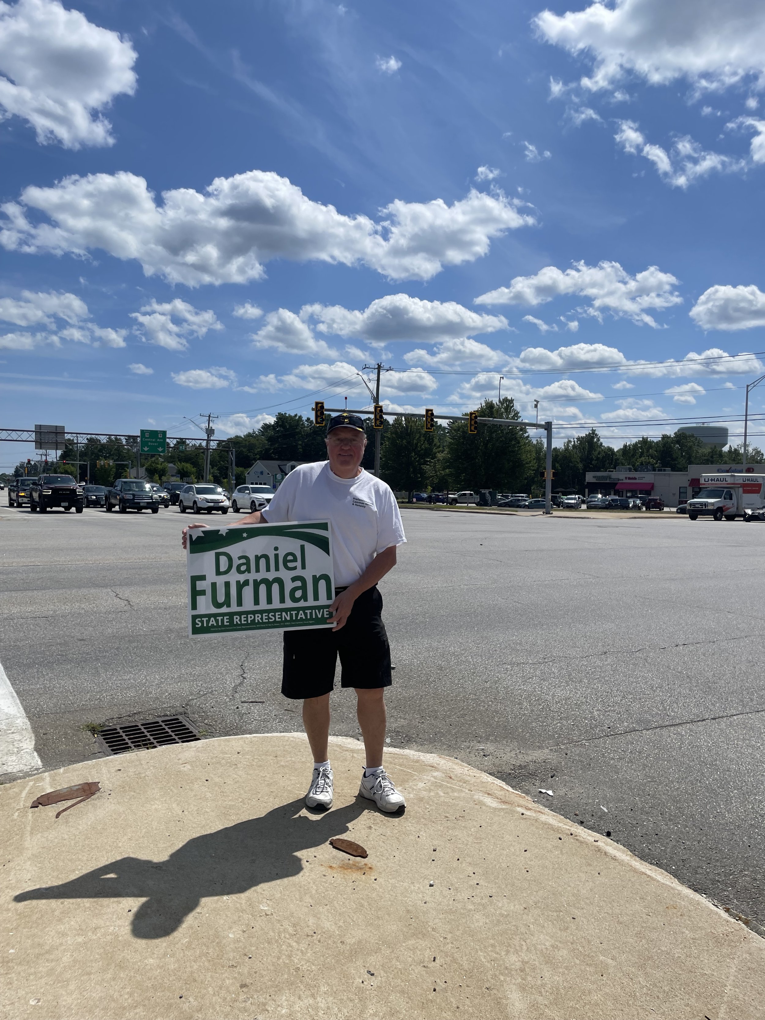 A man holding a sign with the name Daniel Furman in front of a busy intersection with traffic lights and cars. The sky is partly cloudy.