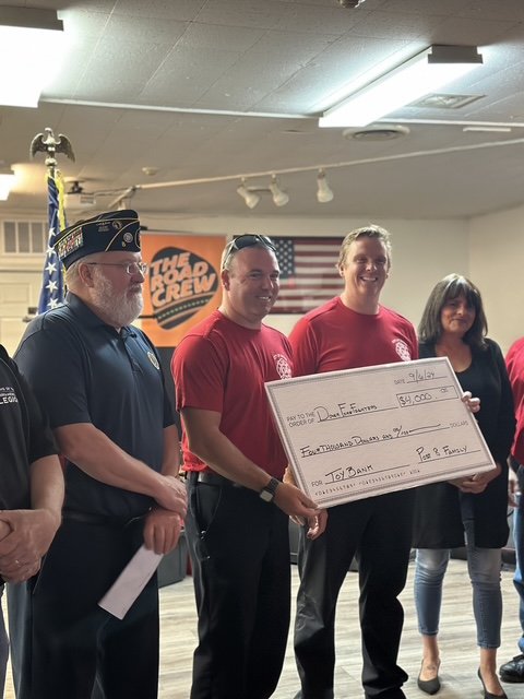 Group of people at an event, with one person holding a large check, American flags, and a banner in the background.