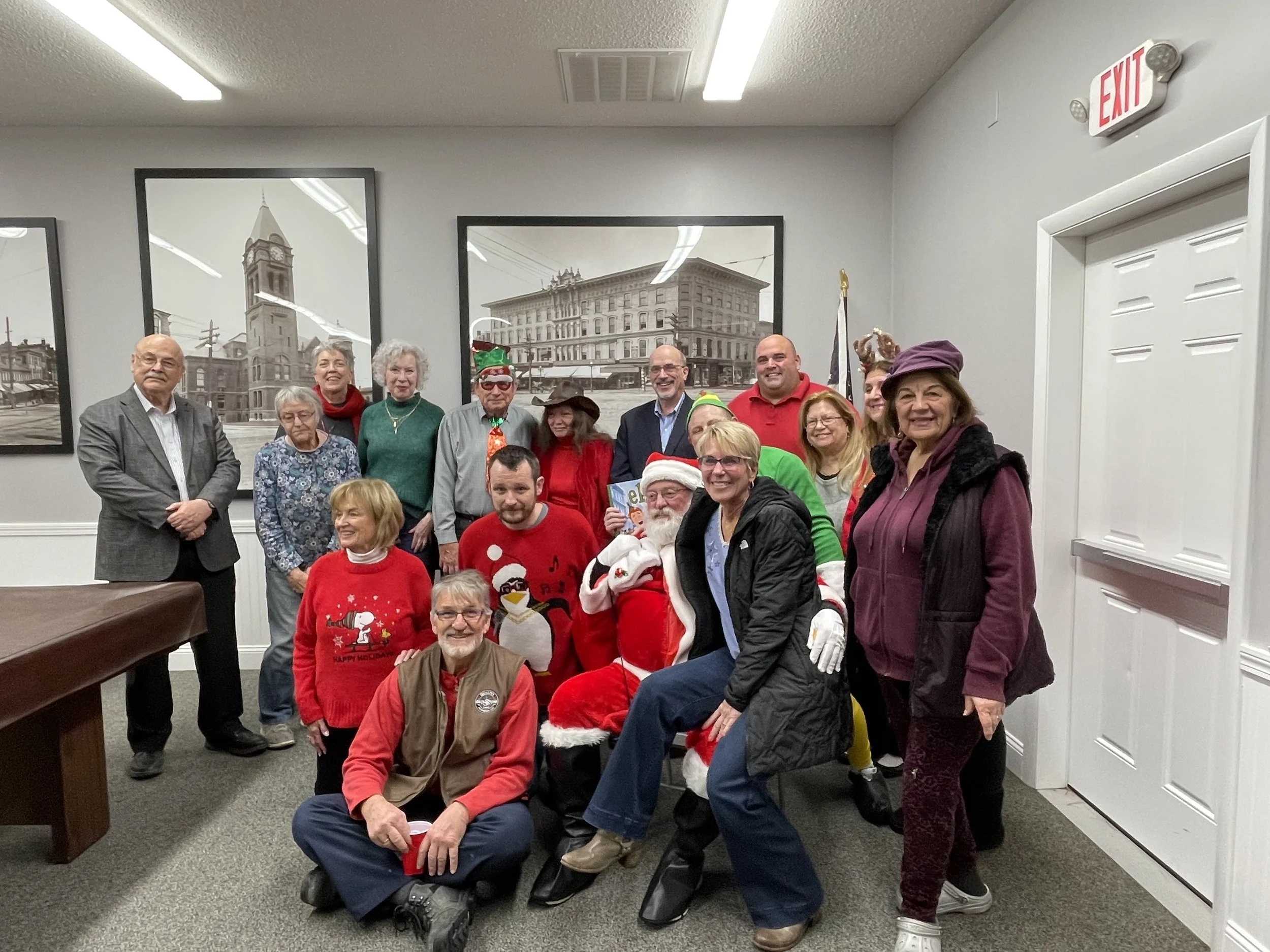 A group of people gathered indoors, some wearing Christmas sweaters and holiday accessories, with a few individuals dressed as Santa Claus and elves. The background features framed black-and-white photos of buildings and a white wall with an exit sign.