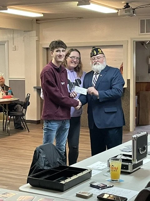 A young man and a woman shaking hands with an older man in a military hat, in a room with tables, chairs, and a PowerPoint projector.
