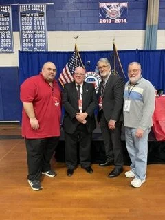 Four men standing together in a gymnasium in front of a United States flag and a blue banner with sports team banners, posing for a photo.