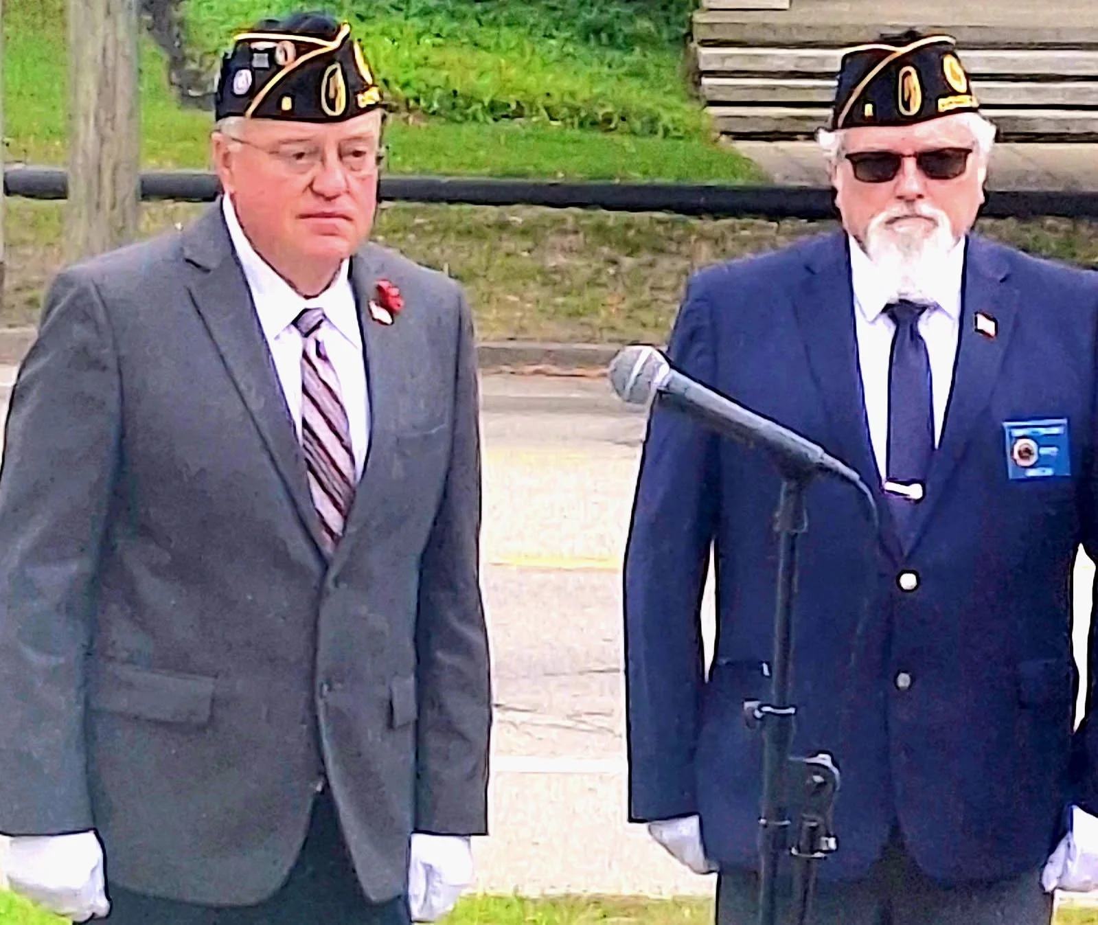 Two men dressed in suits and wearing military-style hats with gold insignia, standing outdoors near a microphone, with a wooden staircase and trees in the background.