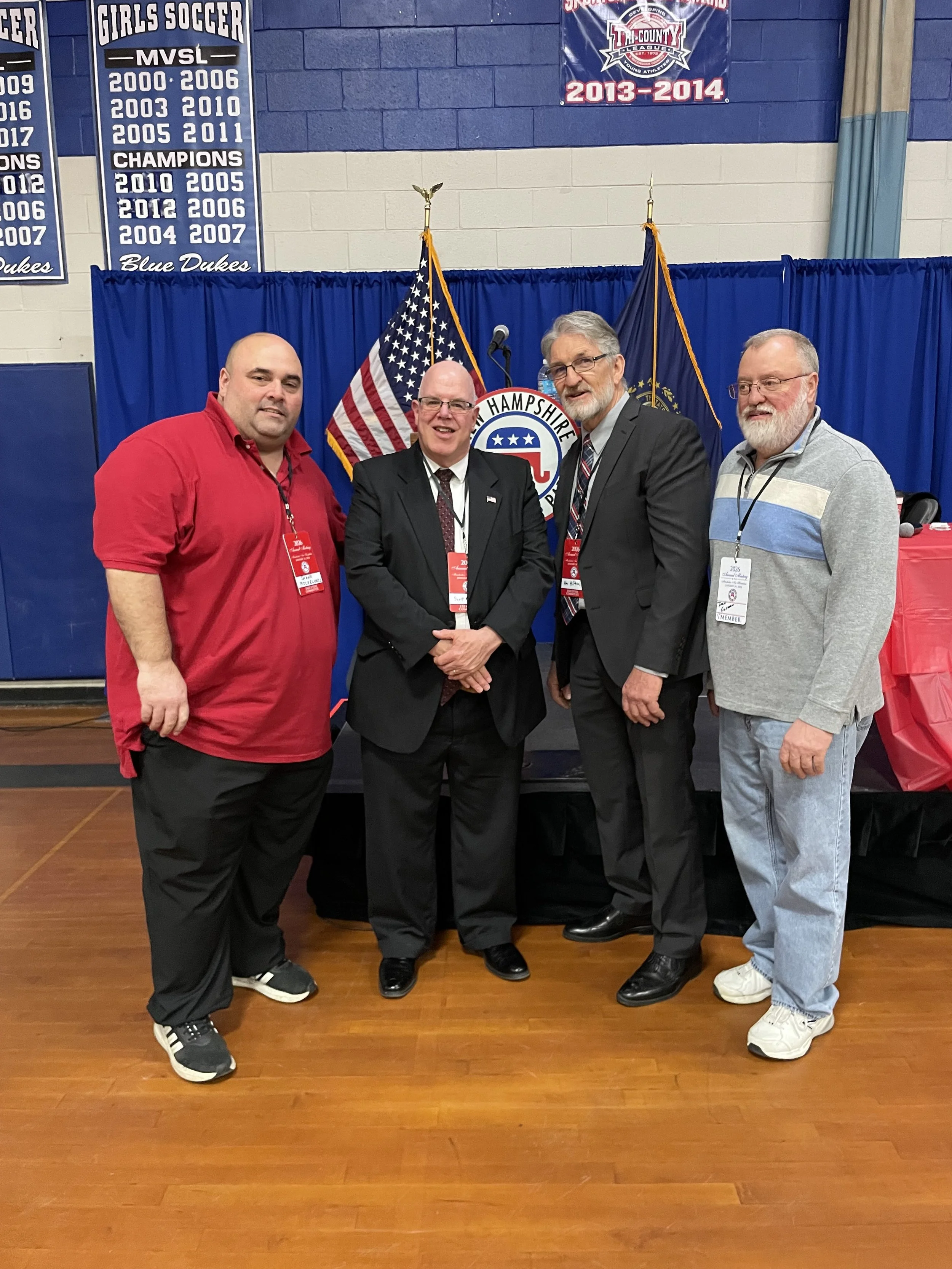 Four men standing in front of blue curtains with American and state flags behind them. They are at a political event in New Hampshire, as indicated by the badge and banners on the wall.