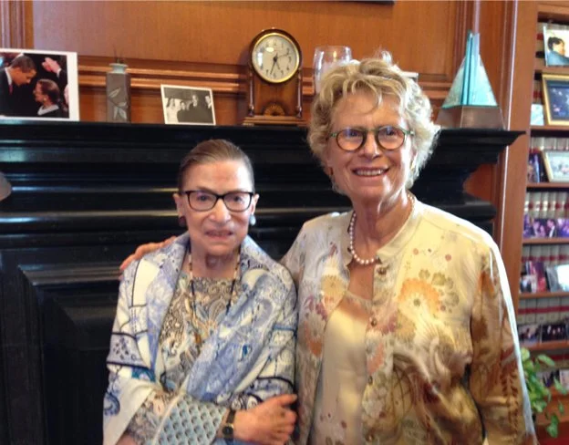 Two smiling women standing indoors in front of a black fireplace with photos and a clock on the mantel. One woman is wearing glasses, a patterned blouse, and a shawl, while the other is wearing glasses, a floral jacket, and a pearl necklace.