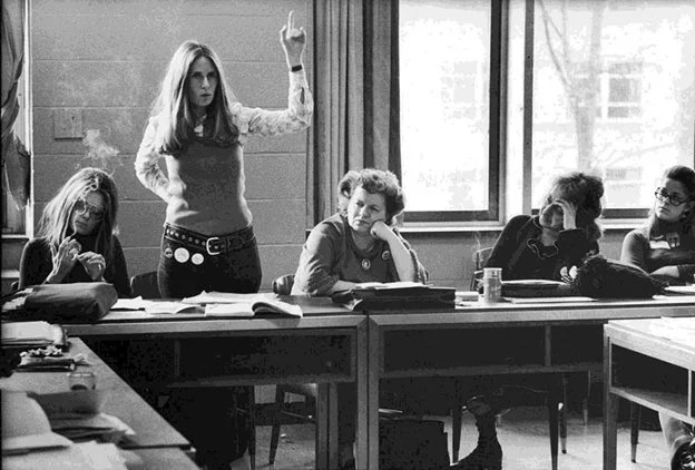A woman is standing at a desk in a classroom, raising her hand, with four women seated at the desk, appearing to listen.