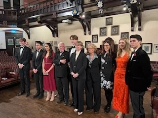 A group of nine people dressed formally, standing in a line, inside a room with wooden beams and framed pictures on the wall.