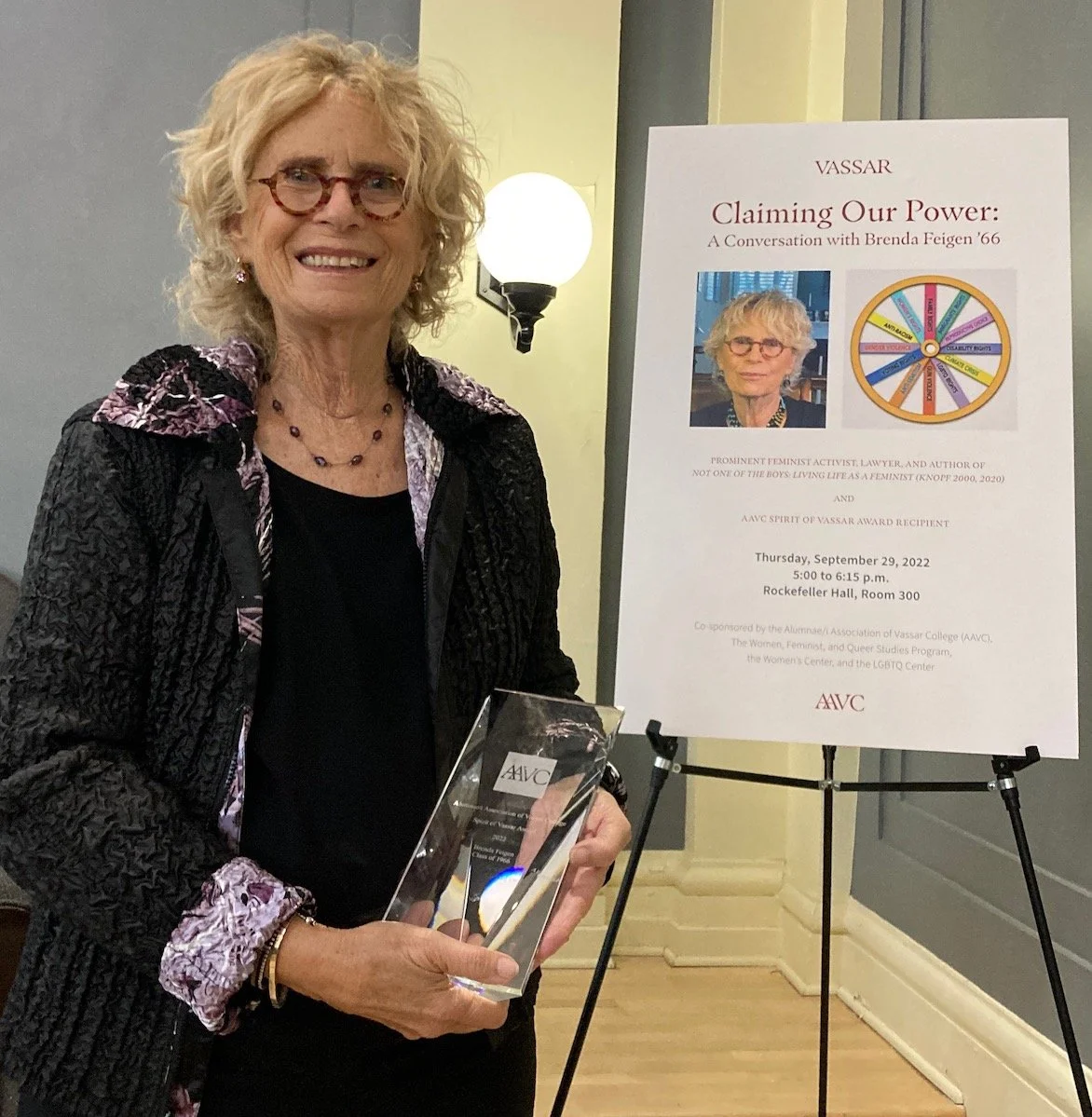 A woman with curly blonde hair, glasses, and a black quilted jacket holding a clear award, standing next to a poster about an event titled 'Claiming Our Power' with her photo and event details.
