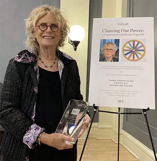 An older woman with curly blonde hair, glasses, and a dark jacket holding a clear award, standing next to a poster about a talk on empowerment and claiming power, dated September 29, 2022.