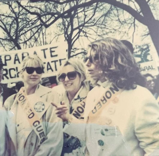 Three women with sunglasses and sashes participating in a protest or march outdoors with trees in the background and a sign in the distance.