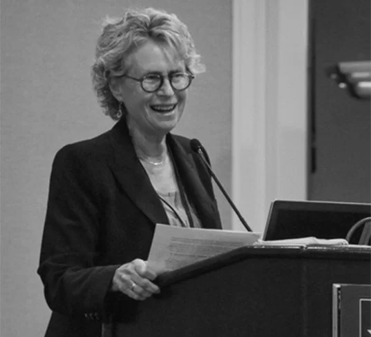 An older woman with curly gray hair and glasses speaking at a podium with a microphone, smiling, in a professional setting.