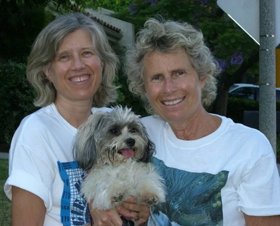 Two women smiling outdoors, holding a small fluffy dog.