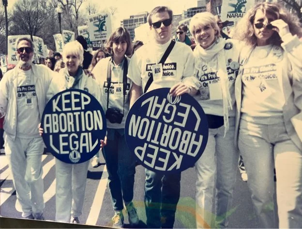 Group of people participating in a pro-choice rally, holding signs that read 'Keep Abortion Legal' with a large crowd and protest banners in the background.