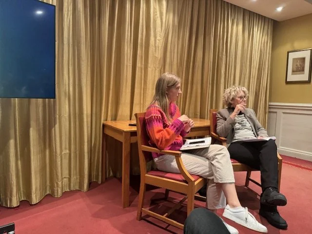 Two women are seated and having a conversation in a room with gold curtains and framed pictures on the wall.