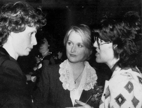 Three women engaged in conversation at a social gathering, with two women facing the camera and one woman facing away.