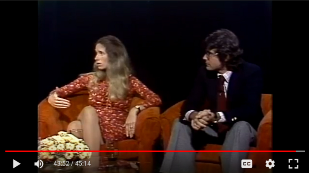 A woman with long, wavy brown hair wearing a red floral dress is sitting on an orange sofa. A man with dark, curly hair and glasses, dressed in a dark blazer, white shirt, and gray pants, is sitting next to her. They are engaged in conversation on a talk show set with a dark background.