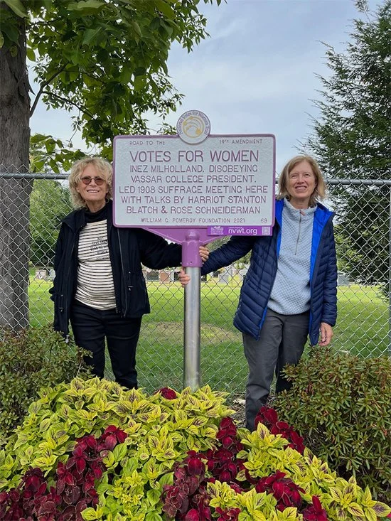 Two women standing on either side of a historical marker about women's voting rights, surrounded by flowering plants and a chain-link fence in the background.