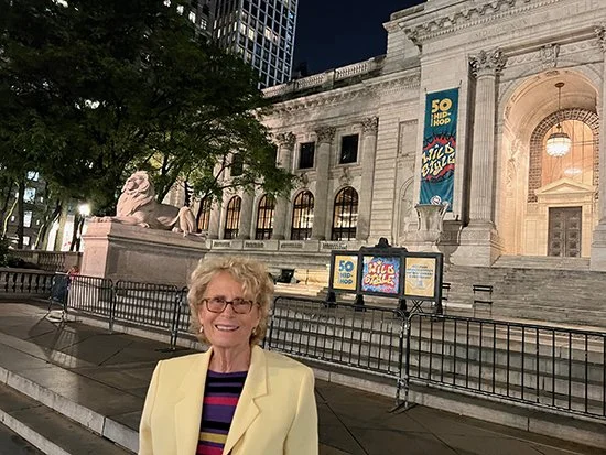 An older woman with curly blonde hair and glasses smiling in front of a historic building with marble columns and lion statues, lit up at night, with banners advertising a 50th anniversary celebration.