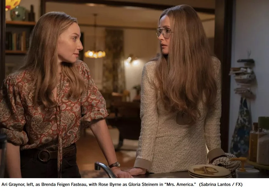 Two women having a serious conversation in a cozy kitchen. The woman on the left has long, light brown hair and wears a patterned blouse. The woman on the right has long, wavy hair, glasses, and wears a beige blazer. The kitchen has warm lighting, books, and kitchenware.