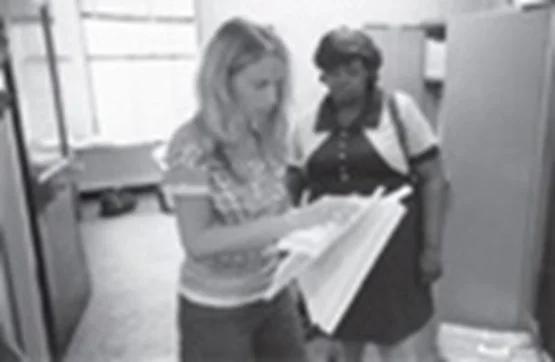 Two women standing in an office, looking at a large document or book together.