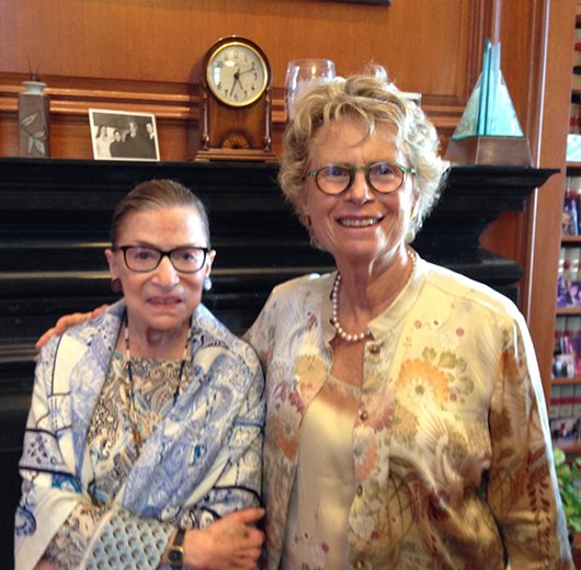Two women standing close together indoors, smiling at the camera. One woman wears glasses and a patterned blouse, the other also wears glasses and a floral jacket. Background shows a fireplace, a clock, and a storefront window.