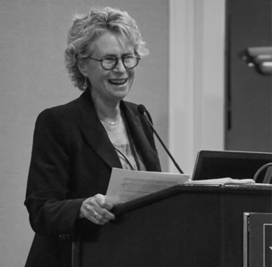 A woman with curly hair and glasses smiling while speaking at a podium with a microphone, holding a paper in her hand