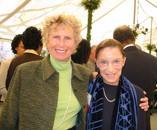 Two women smiling at a social event, one with curly gray hair wearing a green turtleneck and a blazer, and the other with glasses and a blue scarf, with people in the background under a tent.