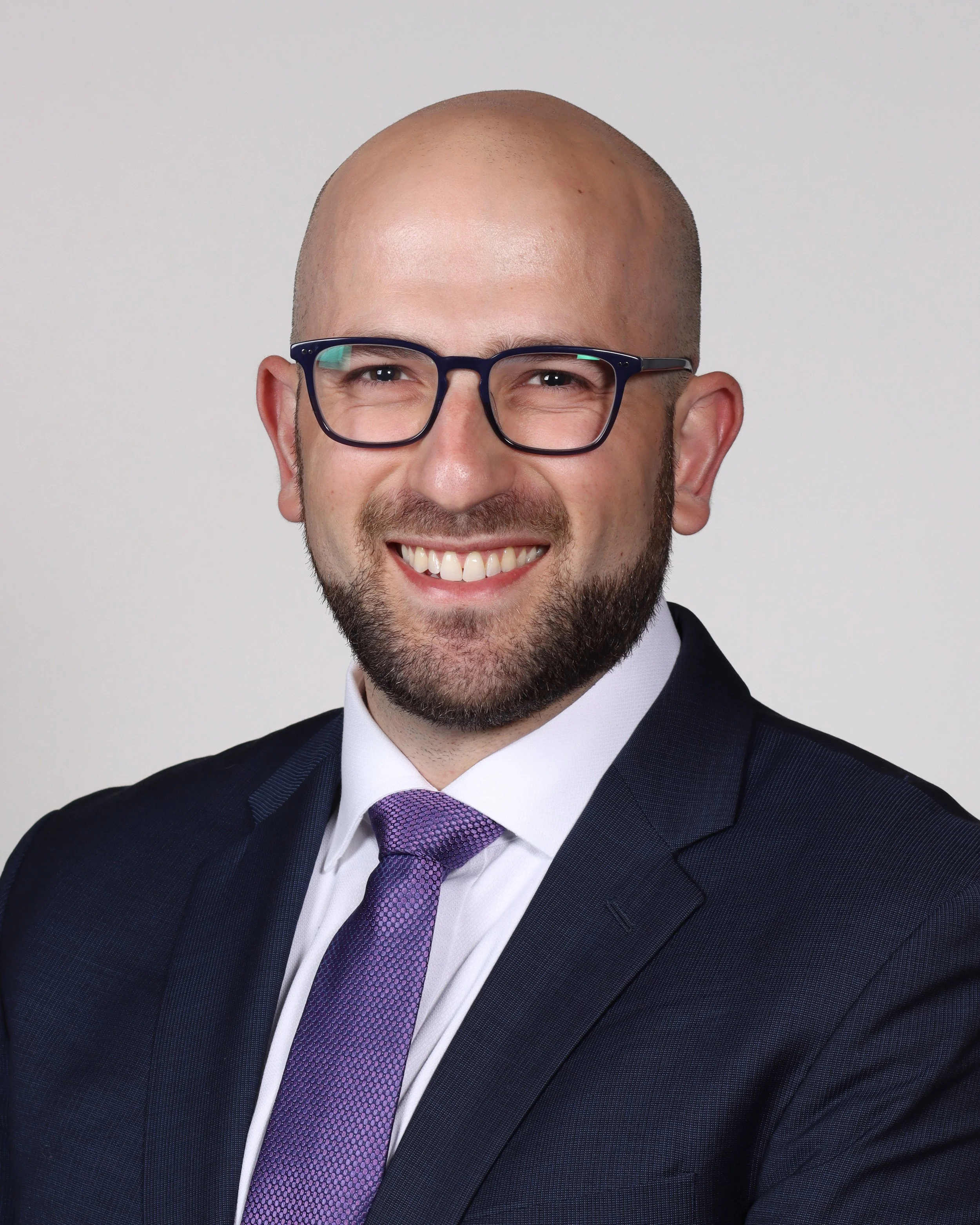 Portrait of Dr. Ari Bernstein, New Jersey urologist, a smiling man wearing glasses, a dark suit, white shirt, and purple tie, with a shaved head and beard, against a plain light background.