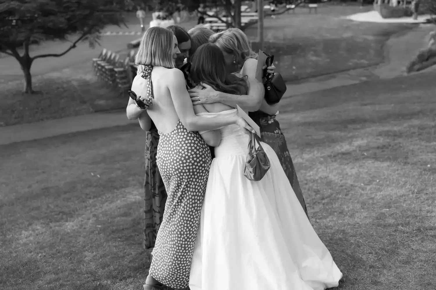 A group of women embracing and hugging each other outdoors on a grassy area, with trees and benches in the background.