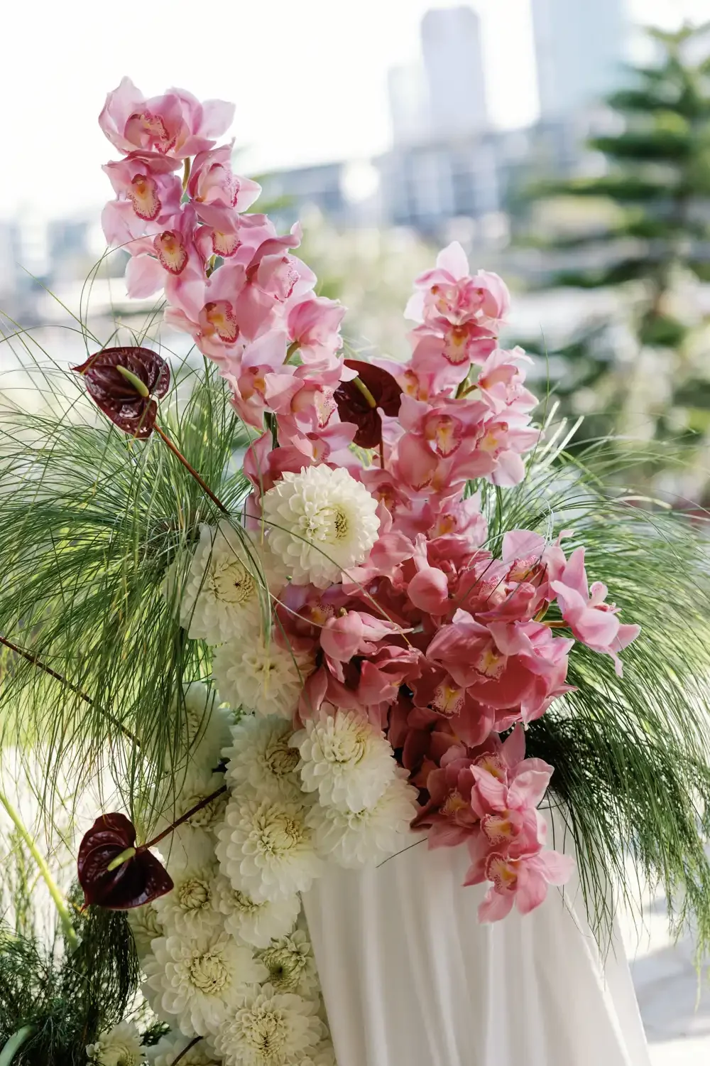 A vibrant floral arrangement with pink orchids, white dahlias, and green foliage in a white vase, with an urban cityscape background.