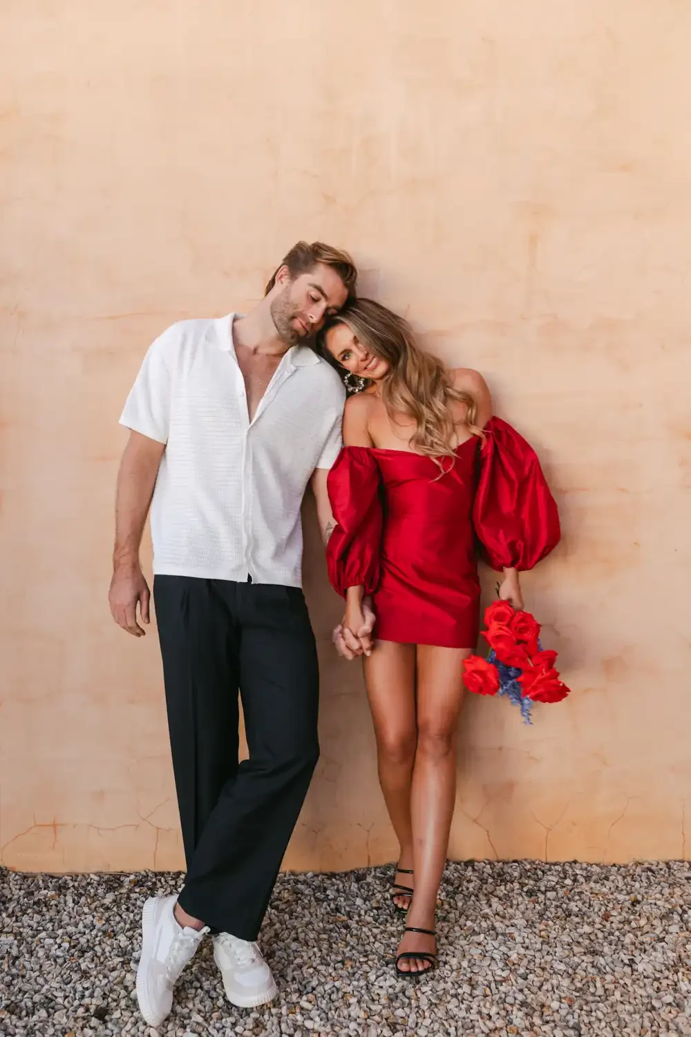 A man and woman standing close together against a plain beige wall, holding hands. The man wears a white short-sleeve shirt and black pants, while the woman is in a red off-shoulder dress with puffed sleeves, black strappy heels, and holds a bouquet 