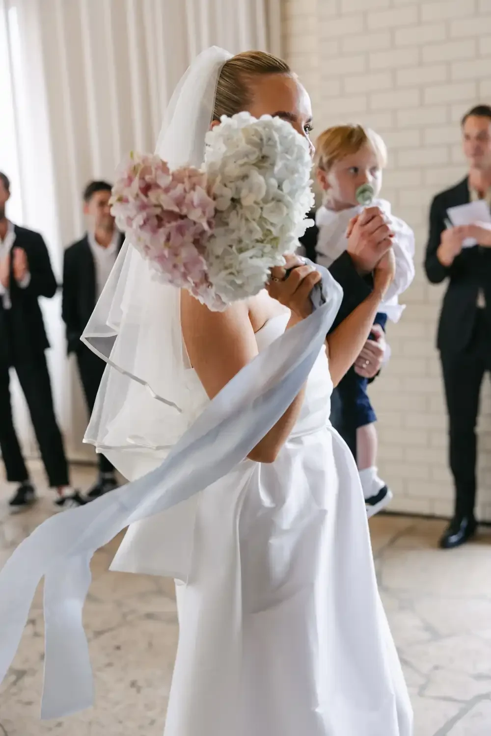 Bride holding a bouquet of pink and white flowers, with a bride veil, in a wedding ceremony. Children and adults are in the background.